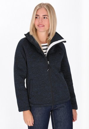 Nuori nainen, joka käyttää tummansinistä vetoketjullista fleece-takkia valkoisella sherpa-vuorella, sinisiä farkkuja ja raidallista paitaa, katsoo oikealle ja hymyilee hieman.