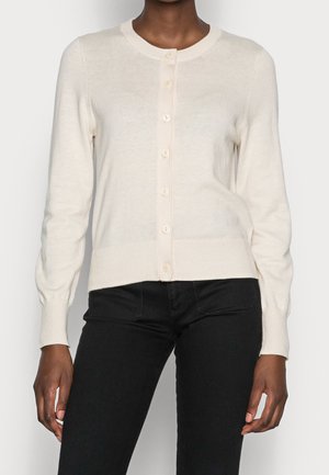 Frau trägt eine cremefarbene, geknöpfte Strickjacke und schwarze, eng anliegende Hose, steht vor einem schlichten hellen Hintergrund.