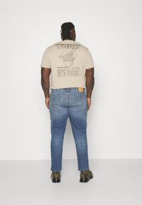 Beige grafisk t-skjorte med trykt nudeldesign på baksiden, kombinert med lyseblå denimjeans og mørkegrønne sko.