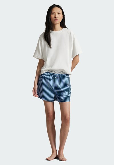 Kvinna som står barfota klädd i en lös vit t-shirt och blårandiga boxershorts med en synlig midja från "Polo Ralph Lauren".