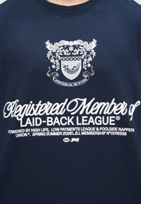 Μπλε πουκάμισο ναυτικού με λευκό κείμενο που αναγράφει «Εγγεγραμμένο Μέλος της Φιλικής Λίγκας», με έμβλημα όπου απεικονίζεται ένας άνθρωπος που χαλαρώνει σε αιώρα πάνω από το κείμενο.