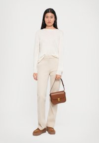 Femme portant un pantalon beige, un pull blanc, tenant un sac à main en cuir marron, se tenant contre un fond blanc uni.