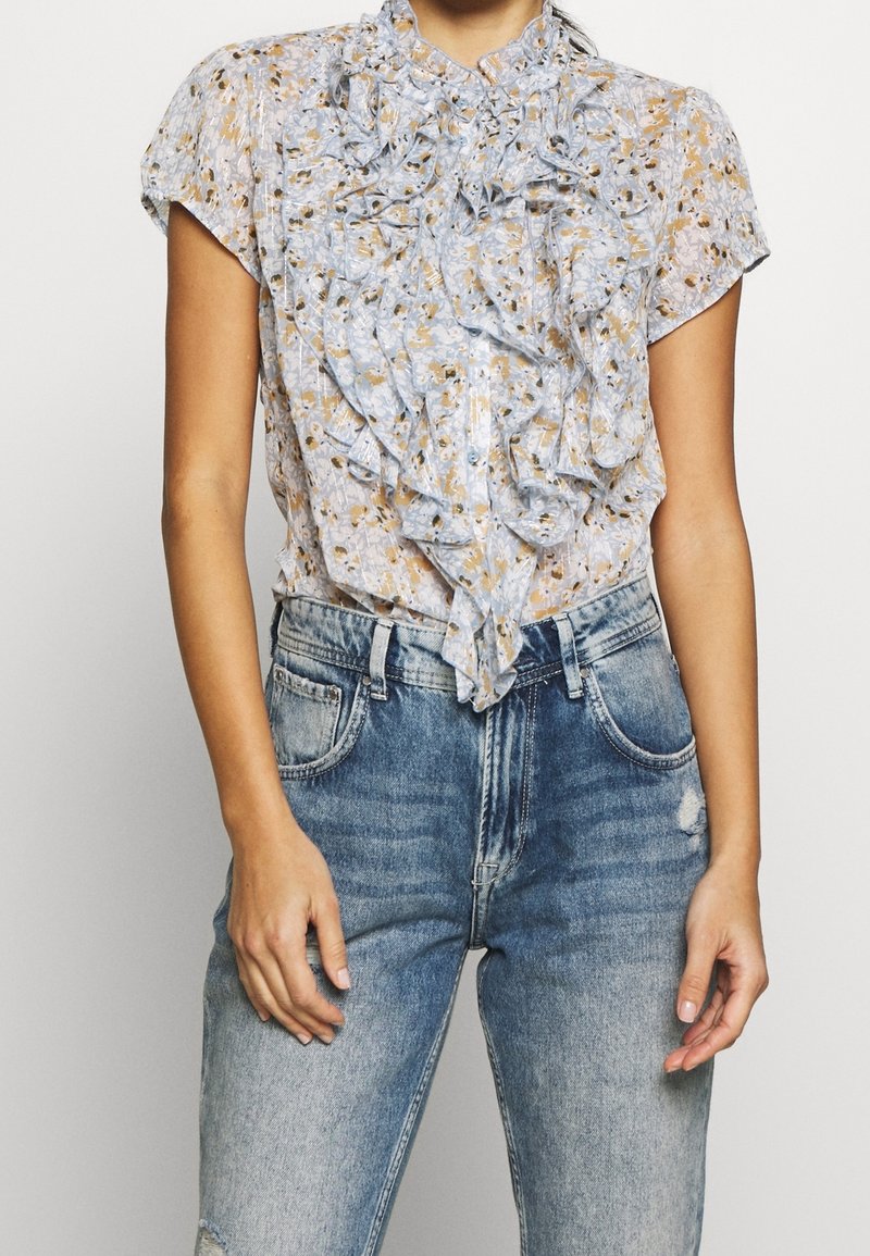 Blusa floral de color azul claro con volante en el frente, mangas cortas y cuello alto, combinada con jeans de tiro alto en azul desgastado.