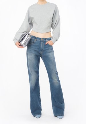 Persoon draagt een lichtgrijze cropped sweatshirt, hoog getailleerde wijde blauwe jeans en houdt een zilveren clutch vast tegen een witte achtergrond.