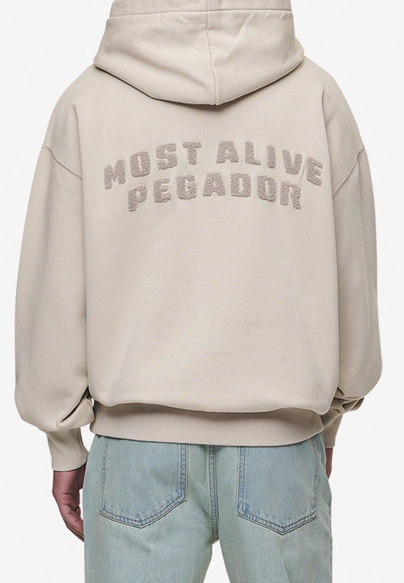 Personne portant un sweat à capuche beige avec le texte texturé "MOST ALIVE PEGADOR" dans le dos et un jean bleu clair, vu de derrière.