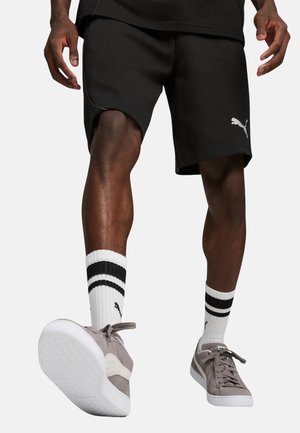 Uomo che indossa pantaloncini neri Puma, calzini bianchi con strisce nere e scarpe da ginnastica grigie Puma, in piedi con un piede leggermente sollevato.