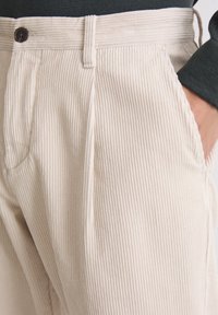 Pantalon en velours côtelé beige avec des côtes verticales, doté d'une fermeture à bouton, de poches latérales et de jambes légèrement fuselées pour un ajustement près du corps.