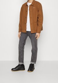 Brown cotton jacket with a button front and two pockets, worn over a white t-shirt and grey jeans, paired with black sneakers featuring gum soles.