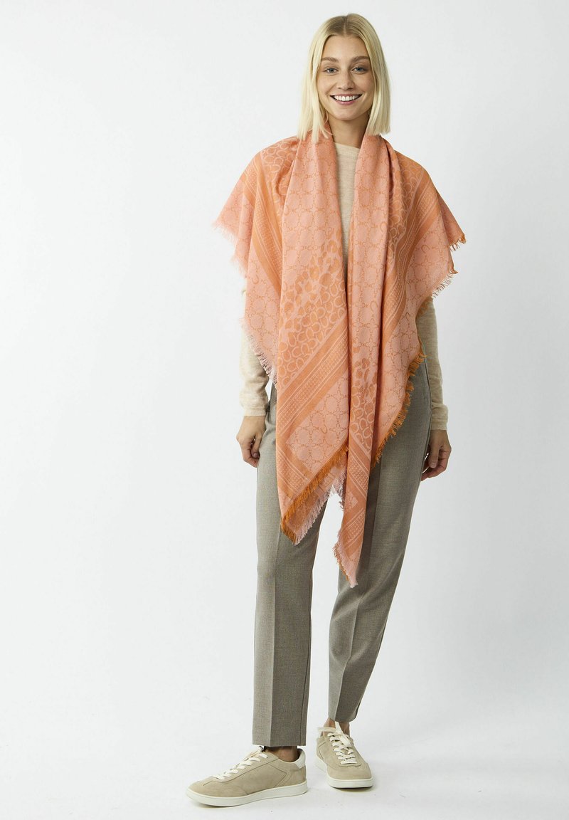 Aprikosenfarbener Schal mit Fransen, der ein geometrisches Muster und gestreifte Designelemente aufweist, getragen über einem hellen Oberteil und einer Hose.