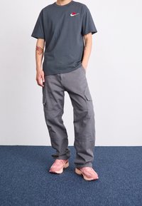 Szara bawełniana koszulka z małym haftowanym różą i logo Nike, w połączeniu z szarymi spodniami cargo i zakurzonymi różowymi butami sportowymi.