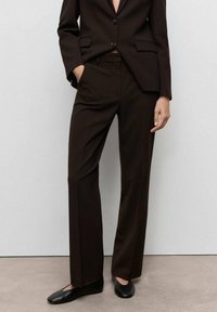 Brown tailored suit pants with a flat front, straight legs, and subtle crease detailing. Worn with fitted black shoes.