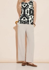 Woman wearing sleeveless black and white patterned top, beige wide-leg pants, brown slip-on shoes, and chunky bracelets, hands in pockets.