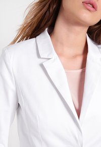 Biały żakiet z wąskim klapem, gładką teksturą i delikatnym połyskiem, założony na miękki różowy top z okrągłym dekoltem.