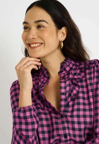 Blusa a cuadros rosa y azul marino con cuello con volantes, cierre de botones en la parte delantera y detalles de puños fruncidos. Textura de tela suave. Pendientes de aro de oro.