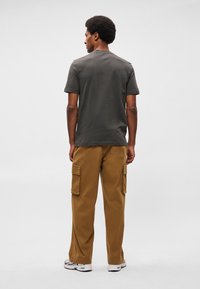 Pelēks kokvilnas T-krekls ar apaļu kaklu, apvienots ar brūnām kravas biksēm, kas raksturīgas ar divām sānu kabatām. Stils ir brīvs, minimāla dizaina.