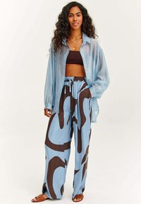 Svietloj modrá priehľadná košeľa na gombíky, hnedý crop top a voľné modré nohavice s veľkými hnedými abstraktnými vzormi; v kombinácii s hnedými sandálmi.