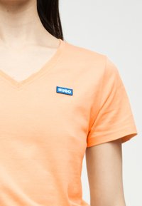 Κοντινό πλάνο ενός ατόμου που φοράει ροδακινί μπλουζάκι με V λαιμόκοψη και ένα μικρό μπλε ορθογώνιο σήμα με την επιγραφή "HUGO" στο αριστερό μέρος του στήθους.