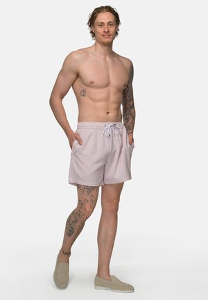 Oberkörperfreier Mann mit Tätowierungen an Arm und Bein, der hellrosa Shorts mit Kordelzug und beige Slip-On-Schuhe trägt und mit den Händen in den Taschen steht.