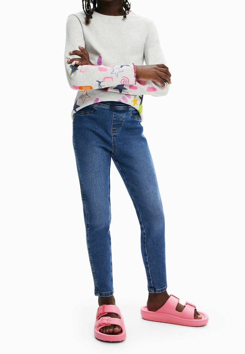 Chemise à manches longues gris clair avec des motifs colorés, associée à un jean skinny bleu et des sandales en caoutchouc rose avec deux sangles réglables.