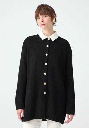Black ribbed cardigan with a white collar, featuring large gold buttons down the front. The fabric appears soft with a relaxed fit.