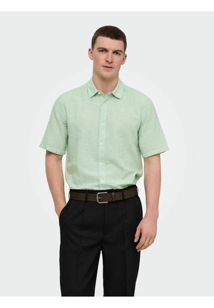 Mann trägt ein hellgrünes, kurzärmeliges Hemd mit Knöpfen, in schwarze Hosen mit einem braunen Gürtel gesteckt, steht mit einer Hand in der Tasche.