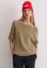 Beige gebreide trui met een losse pasvorm en brede halslijn, gecombineerd met zwarte broek. Een rode geribbelde beanie ligt op lang, steil haar.