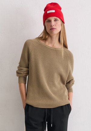 Beige gestrickter Pullover mit lockerer Passform und weiter Ausschnitt, kombiniert mit schwarzen Hosen. Eine rote gerippte Mütze sitzt auf langen, glatten Haaren.