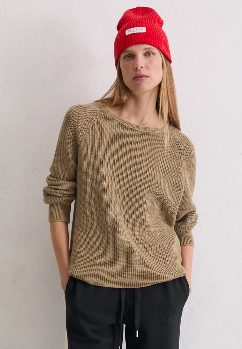 Beige gebreide trui met een losse pasvorm en brede halslijn, gecombineerd met zwarte broek. Een rode geribbelde beanie ligt op lang, steil haar.