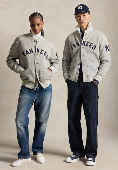 Deux mannequins portent des vestes grises des Yankees et des jeans, l'un avec des baskets blanches, l'autre avec des baskets bleues et une casquette des Yankees.