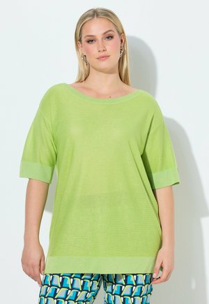 Lyshåret kvinde iført en løs, lysegrøn top med korte ærmer og bukser med geometrisk mønster i blå, grøn og hvid, stående mod en hvid væg.