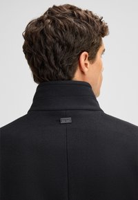 Cappotto in misto lana nero con colletto alto e un'etichetta sul retro. Il tessuto sembra morbido e testurizzato, adatto per le giornate fresche.