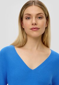 Blauer Strickpullover mit V-Ausschnitt-Design, der ein strukturiertes Muster und eine lockere Passform bietet, die das detaillierte Gewebe des Stoffes hervorhebt.