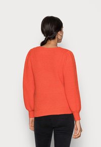 Maglione di lana arancione brillante con una vestibilità comoda, caratterizzato da una texture a coste, scollo rotondo e maniche a palloncino. Indossato con jeans neri.