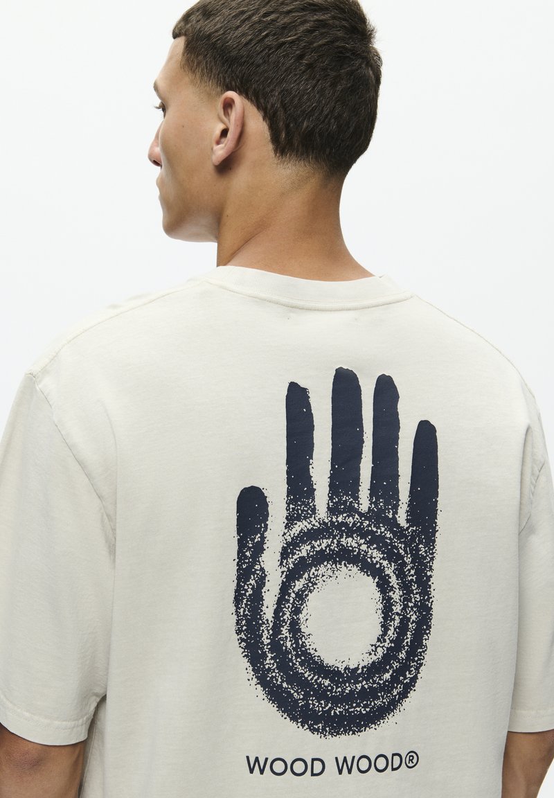 T-shirt en coton couleur crème avec un grand motif de main navy au dos, accompagné de détails circulaires et du texte 'WOOD WOOD®'.