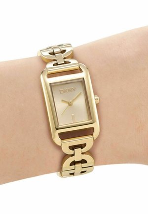 Goudkleurig DKNY polshorloge met rechthoekige wijzerplaat en schakelsarmband om de pols van een persoon, met uur-, minuut- en secondewijzers.
