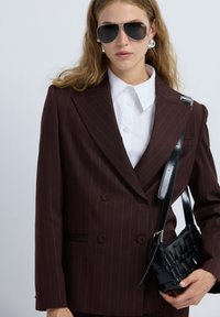 Mujer vistiendo un blazer de rayas oscuras de doble botonadura, camisa blanca con cuello, grandes pendientes de plata, gafas de sol oscuras y sosteniendo un bolso de mano negro de mimbre.