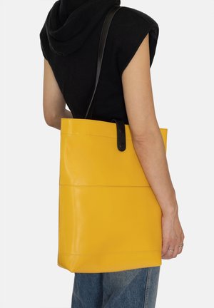 Gul læder tote taske med en glat tekstur, prydet med et sort håndtag og en knaplukning. Minimalistisk design med horisontale sømdetaljer.