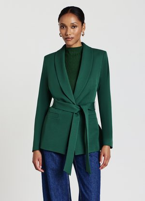 Blazer verde scuro con colletto a scialle, cintura da annodare e due tasche frontali. Indossato sopra un maglione verde a coste e abbinato a jeans blu.