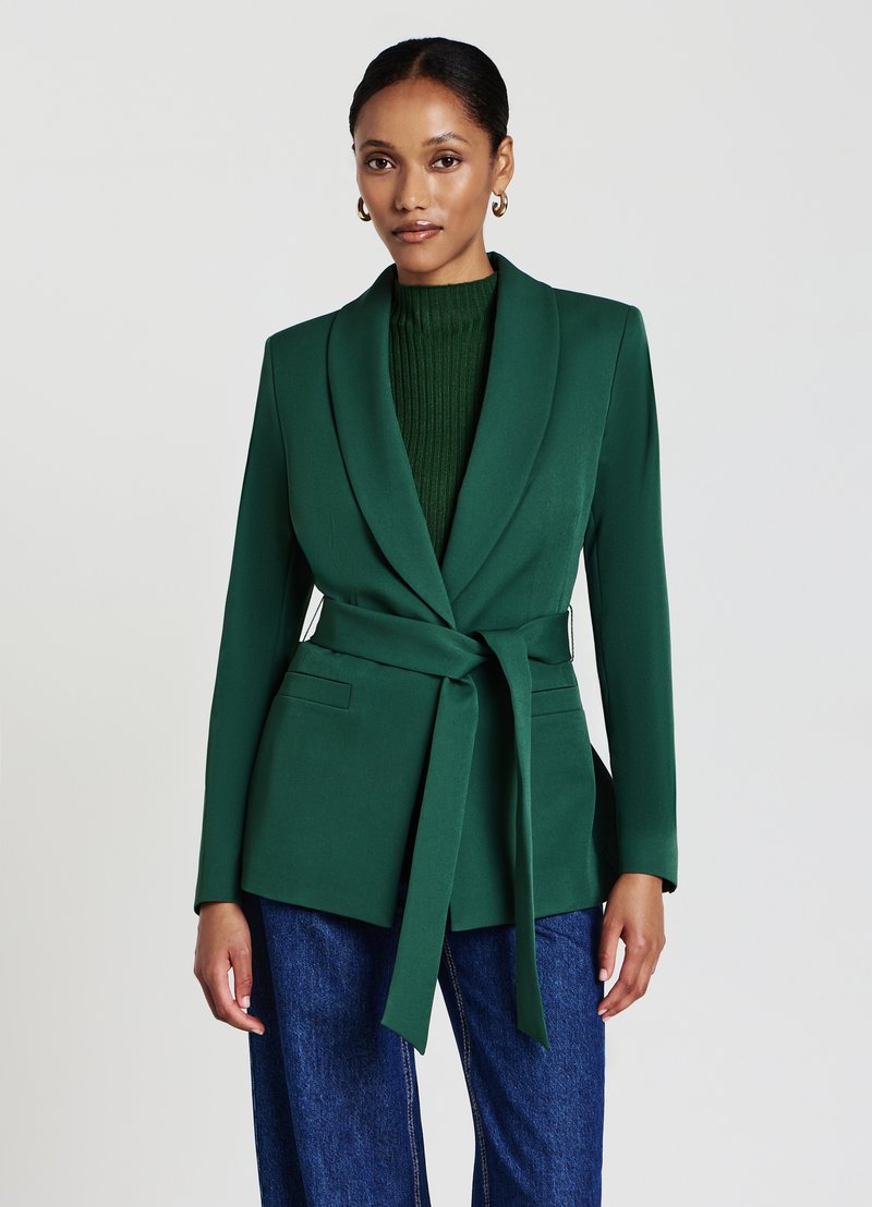 Blazer verde scuro con colletto a scialle, cintura da annodare e due tasche frontali. Indossato sopra un maglione verde a coste e abbinato a jeans blu.