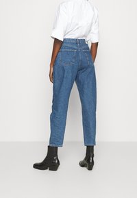 Jeans di denim blu a vita alta con una vestibilità rilassata, dotati di tasche posteriori e design a gamba affusolata. Abbinati a stivaletti neri alla caviglia.