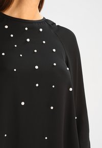 Top nero con tessuto morbido, scollo rotondo, maniche lunghe e perle decorative di diverse dimensioni sparse sul davanti.