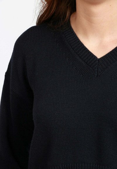 Primo piano di una persona che indossa un maglione a V nero lavorato a maglia con cuciture in evidenza e capelli castani sulle spalle.