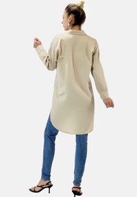 Beige långärmad skjorta med krage, rundad nederkant och avslappnad passform. I kombination med blå jeans och svarta remsandaler. Enkel design, minimala detaljer.