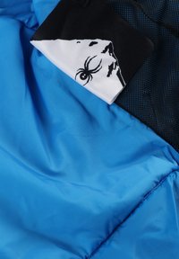 Blauer Stoff mit glatter Textur und matter Oberfläche, ausgestattet mit einem schwarzen Mesh-Akzent und einem weißen Stoffetikett, das eine Grafik einer Spinne zeigt.