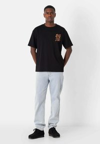 Camiseta de algodón negra con mangas cortas, que presenta un pequeño gráfico naranja en el lado izquierdo del pecho. Combinada con jeans de mezclilla azul claro y zapatos negros.