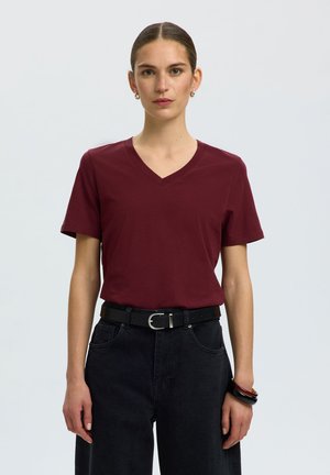 Kvinde med glat hår trukket tilbage iført en vinrød V-hals T-shirt, sort bælte, sorte jeans og sorte og røde armbånd på højre håndled.
