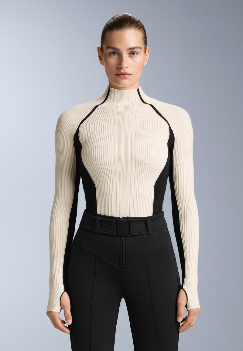 Krémový vysoký top s ribbovaným vzorem a černými bočními panely. Má dlouhé rukávy s otvory na palce a přiléhavý střih, v kombinaci s černými kalhotami.