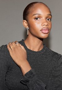 Top nero metallizzato con tessuto strutturato e maniche arricciate. Il modello indossa un anello d'argento e orecchini con forme geometriche.