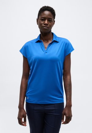 Tievs cilvēks ar īsiem cirtainiem matiem, kurš valkā spilgti zilu īsroku polo krekliņu un tumšas bikses uz vienkārša balta fona.