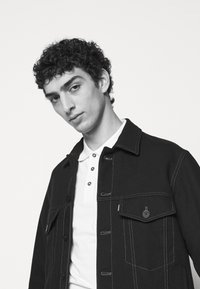 Chaqueta negra con cuello, bolsillos frontales y costuras blancas, llevada sobre una camiseta polo blanca; el modelo tiene el cabello rizado y una expresión neutral.